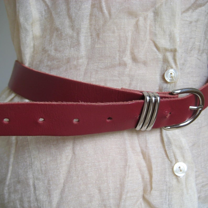 May include: A red leather belt with a silver-colored buckle and loops. The belt is worn over a cream-colored, button-down shirt. The belt has several holes for adjustment.