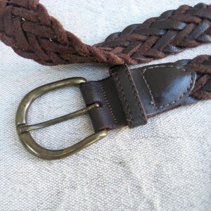 May include: A close-up of a brown braided belt with a brass buckle. The belt is made of woven leather and has a dark brown leather buckle strap. The buckle is a classic oval shape.