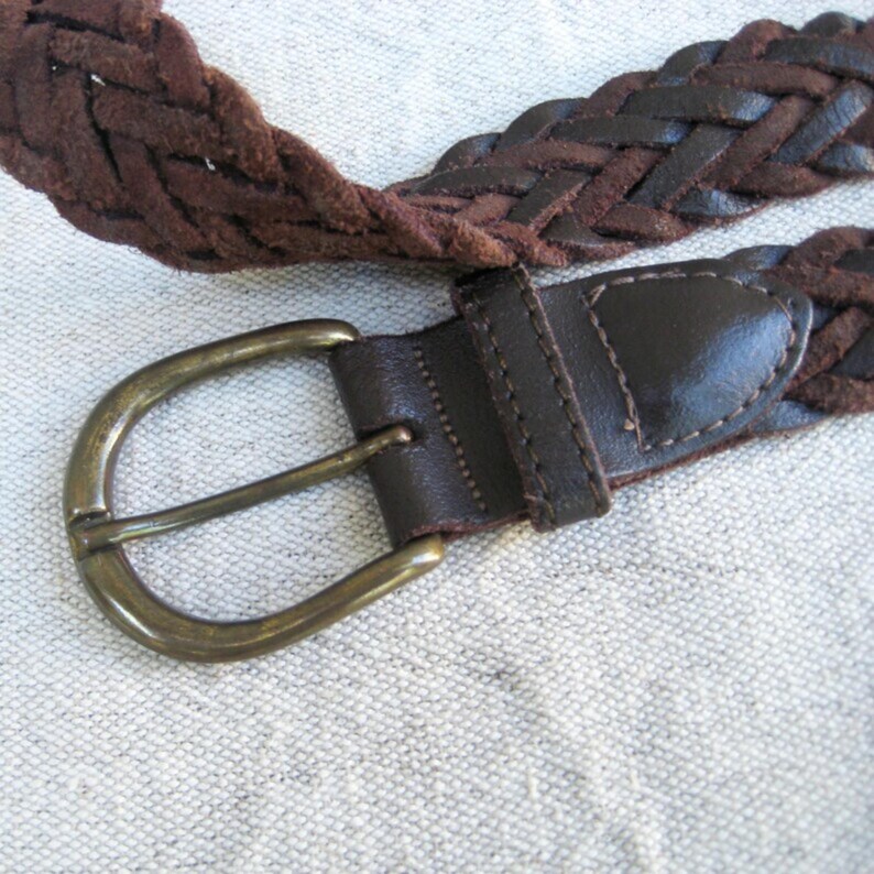 May include: A close-up of a brown braided belt with a brass buckle. The belt is made of woven leather and has a dark brown leather buckle strap. The buckle is a classic oval shape.