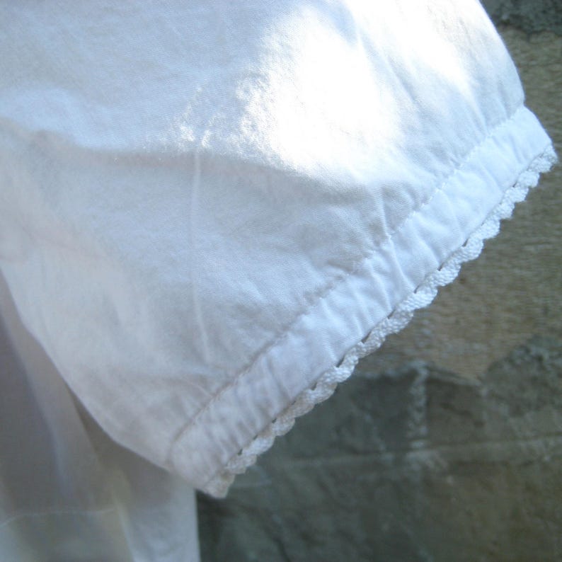May include: Close-up of a white fabric sleeve with a scalloped lace trim. The sleeve is made of a soft, textured material, and the lace trim adds a delicate detail to the edge of the sleeve.