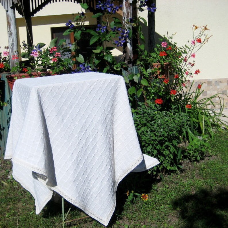 Pu&ograve; includere: Una tovaglia bianca con un motivo a rombi e un bordo in pizzo. La tovaglia &egrave; drappeggiata su un tavolo e posizionata su uno sfondo di piante verdi e fiori.