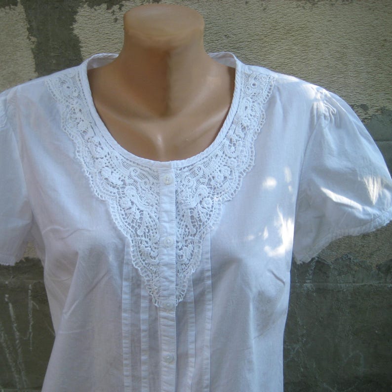 May include: A white short-sleeved blouse with a delicate lace collar and button-down front. The blouse features subtle pleats down the front and a rounded neckline. The fabric appears to be a lightweight cotton or linen.