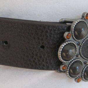 May include: A brown leather belt with a large oval-shaped buckle. The buckle is silver-toned and features a design of black and orange gemstones.