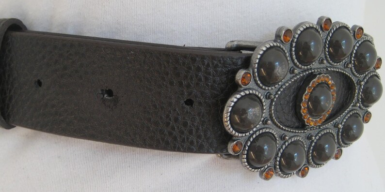 May include: A brown leather belt with a large oval-shaped buckle. The buckle is silver-toned and features a design of black and orange gemstones.