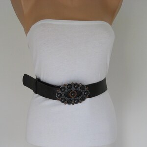 May include: A black leather belt with an oval-shaped metal buckle. The buckle is decorated with a large, dark gray stone surrounded by smaller, copper-colored stones.