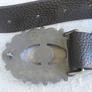 May include: A silver-toned belt buckle with an ornate design featuring a large oval cutout. The buckle is attached to a brown leather belt.