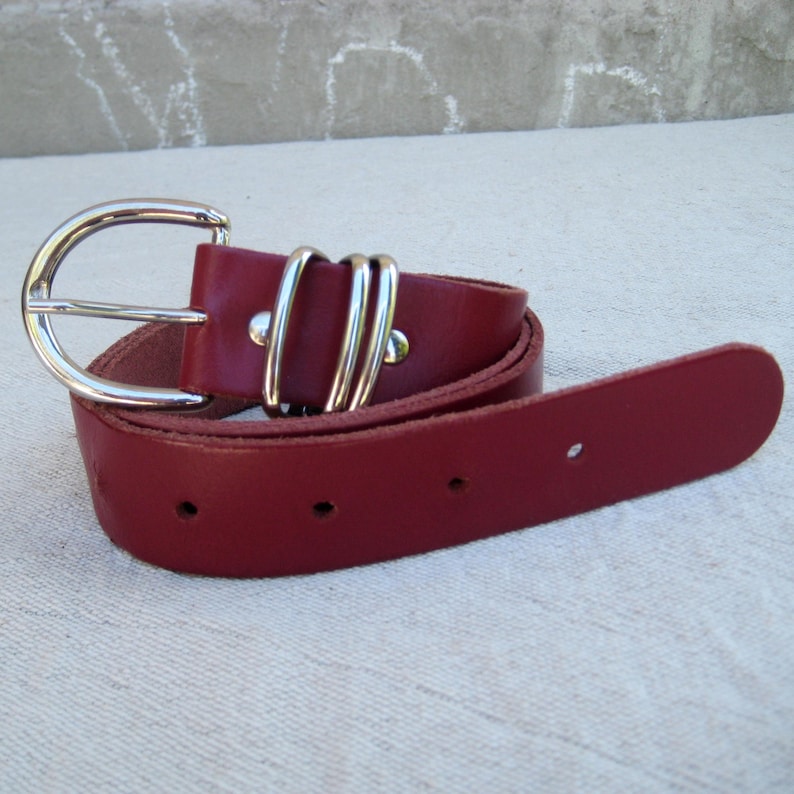 May include: A burgundy leather belt with a silver-colored buckle. The belt has a classic design with a rounded buckle and two silver loops near the buckle. The belt is rolled up on a light-colored surface.