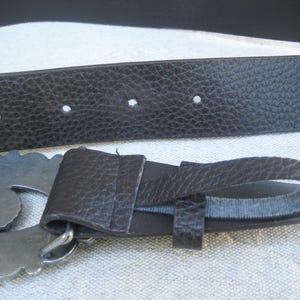 May include: A brown leather belt with a silver buckle featuring a decorative design. The belt has multiple holes for adjusting the fit.