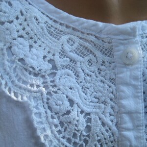 May include: Close-up of a white shirt with intricate, delicate lace detailing around the neckline. The lace features floral and geometric patterns, adding a touch of elegance. A single white button is visible on the shirt's placket.
