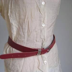May include: A red leather belt with a silver-colored buckle. The belt is worn around a cream-colored blouse. The buckle has a simple, elegant design with three parallel metal bands. The belt has several holes for adjustment.
