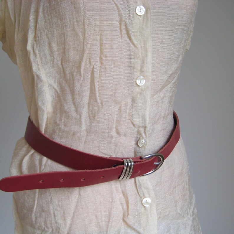 May include: A red leather belt with a silver-colored buckle. The belt is worn around a cream-colored blouse. The buckle has a simple, elegant design with three parallel metal bands. The belt has several holes for adjustment.