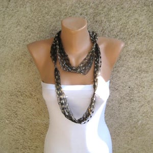 May include: A long, multi-strand necklace featuring a complex weave of thin, metallic-toned threads in shades of black, silver, and bronze. The necklace consists of numerous interconnected loops that create a textured, layered appearance around the neck.