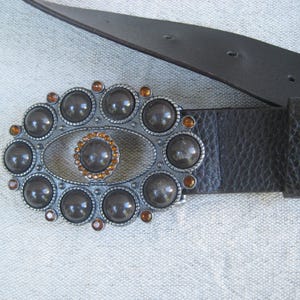 May include: A brown leather belt with a silver buckle featuring a large oval design with a smaller oval in the center. The buckle is decorated with round, dark gray stones and smaller, orange-colored stones.