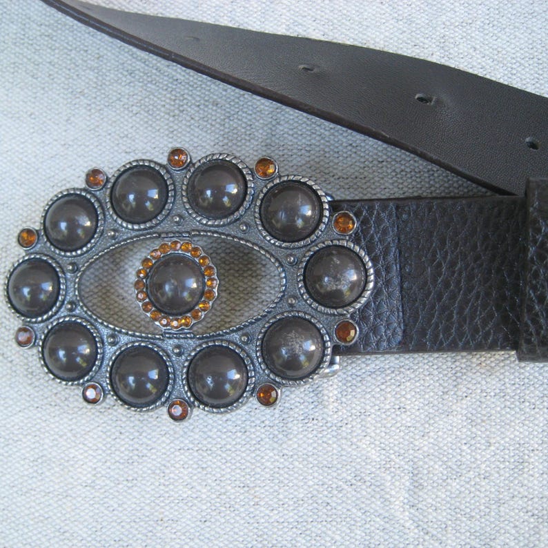 May include: A brown leather belt with a silver buckle featuring a large oval design with a smaller oval in the center. The buckle is decorated with round, dark gray stones and smaller, orange-colored stones.