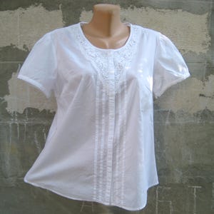 May include: A white short-sleeved blouse with a round neckline and a delicate lace trim. The blouse features vertical pleats down the front and a button-up closure. The sleeves also have a lace trim.