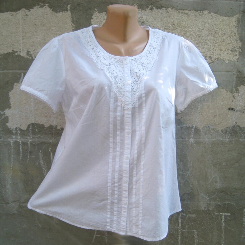 May include: A white short-sleeved blouse with a round neckline and a delicate lace trim. The blouse features vertical pleats down the front and a button-up closure. The sleeves also have a lace trim.