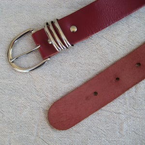 May include: A burgundy leather belt with a silver-colored buckle and three decorative loops. The belt has a classic design with a simple, rounded buckle and several holes for adjustment. The belt is shown on a neutral-colored fabric.