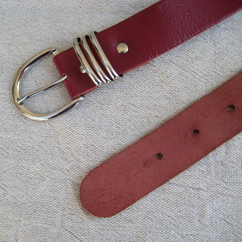 May include: A burgundy leather belt with a silver-colored buckle and three decorative loops. The belt has a classic design with a simple, rounded buckle and several holes for adjustment. The belt is shown on a neutral-colored fabric.