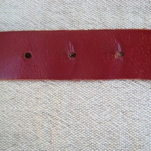 May include: Close-up of a red leather belt with three visible holes. The belt is a solid, deep red color and appears to be made of genuine leather. The holes are evenly spaced and show signs of wear.