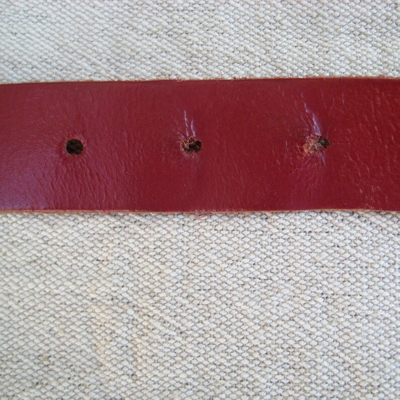 May include: Close-up of a red leather belt with three visible holes. The belt is a solid, deep red color and appears to be made of genuine leather. The holes are evenly spaced and show signs of wear.