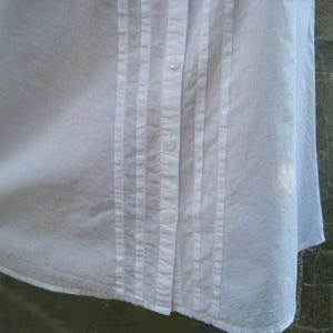 May include: Close-up of a white button-down shirt. The shirt features vertical pleats on the front, running parallel to the button closure. The buttons are small and white, blending with the fabric. The shirt's fabric appears to be a lightweight cotton or linen.