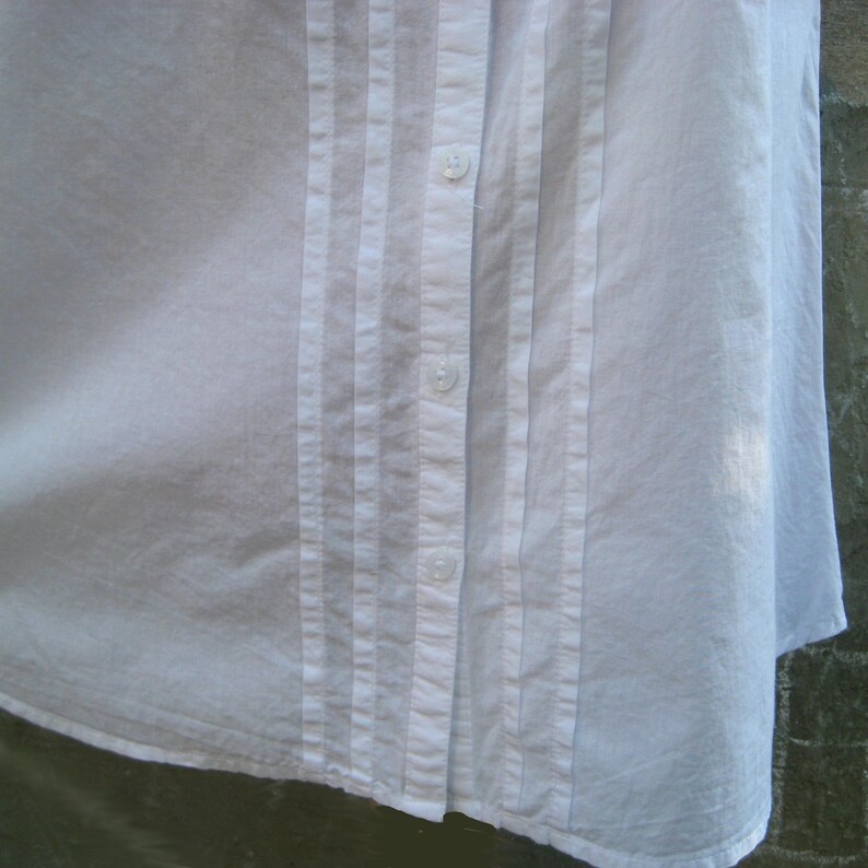 May include: Close-up of a white button-down shirt. The shirt features vertical pleats on the front, running parallel to the button closure. The buttons are small and white, blending with the fabric. The shirt's fabric appears to be a lightweight cotton or linen.