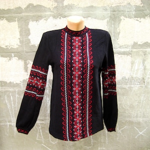 May include: A black long-sleeved sweater featuring a high neckline and intricate red and white geometric embroidery patterns running vertically down the center of the chest and horizontally across the sleeves with ribbed cuffs.