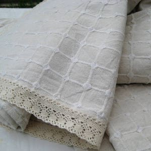 Pu&ograve; includere: Un tovaglia di lino bianco con un delicato motivo ricamato e un bordo di pizzo. La tovaglia &egrave; piegata a met&agrave;, mostrando i dettagli intricati del ricamo.
