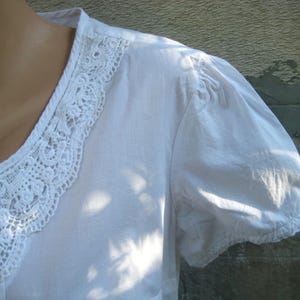 May include: White blouse with a delicate lace trim around the neckline. The short sleeves are gathered at the shoulder and cuff. The fabric appears to be a lightweight cotton or linen. The blouse is a classic, feminine style.