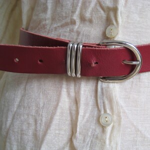 May include: A close-up of a red leather belt with a silver buckle and three silver loops. The belt is worn over a cream-colored shirt. The belt is a classic accessory.