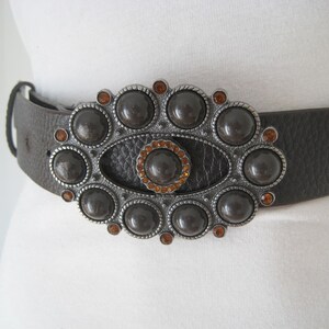 May include: A brown leather belt with a large, ornate silver buckle. The buckle is decorated with a central oval surrounded by a ring of round, dark brown stones. The stones are accented with small, orange rhinestones.