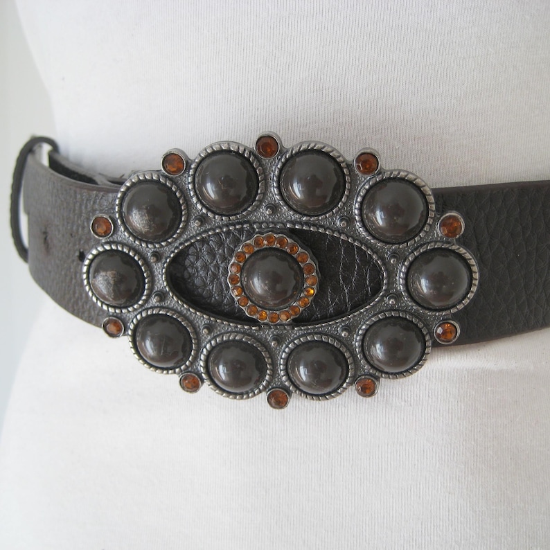 May include: A brown leather belt with a large, ornate silver buckle. The buckle is decorated with a central oval surrounded by a ring of round, dark brown stones. The stones are accented with small, orange rhinestones.