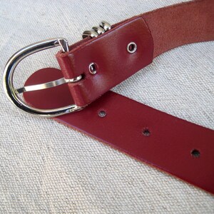 May include: Close-up of a burgundy leather belt with a silver-colored buckle. The belt has a classic design with a rounded buckle and multiple holes for adjustment. The leather appears smooth and durable.