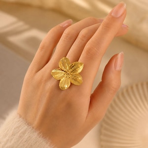 May include: A gold-toned ring featuring a large, five-petaled flower design. The petals have a textured, striated surface finish that radiates from the center of the bloom. The ring is worn on a finger, showcasing the metallic luster and floral shape.