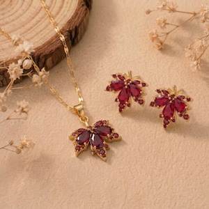 Pode incluir: Um conjunto de colar e brincos a condizer, cada um com um pendente em forma de folha de ácer adornado com pedras vermelho-escuras de corte marquise dispostas em cacho. O colar inclui uma delicada corrente de tom dourado.
