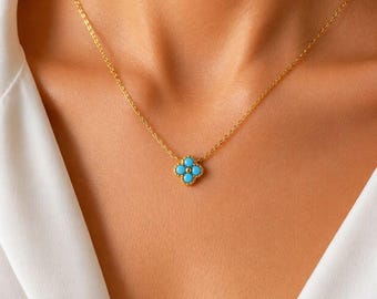 May include: A gold-colored necklace featuring a four-leaf clover pendant with turquoise-colored stones. The necklace is delicate and sits on the neckline of a person wearing a white shirt.