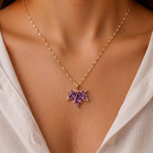 Op de afbeelding: Een goudkleurige ketting met een hanger in de vorm van een esdoornblad. De hanger is versierd met meerdere paarse, peervormige edelstenen in een cluster, geaccentueerd door kleine, heldere, sprankelende steentjes langs de randen van het blad.