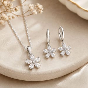 Peut inclure: Un ensemble de bijoux argenté composé d'un collier et d'une paire de boucles d'oreilles créoles. Chaque pièce présente un pendentif ou une breloque en forme de fleur avec cinq pierres transparentes en forme de poire disposées en cercle.
