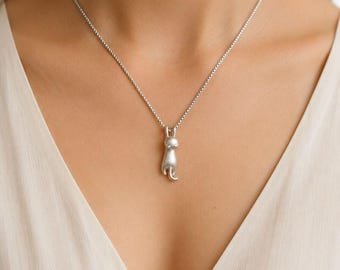 May include: A silver-coloured necklace featuring a pendant shaped like a small, stylised cat hanging by its front paws. The pendant is suspended from a delicate, thin silver-coloured chain that rests against the collarbone area of a white garment.