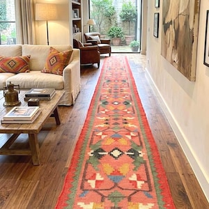 Puede incluir: Una alfombra larga y rectangular con un vibrante fondo coral y un patrón geométrico en tonos verdes, amarillos, azules y blancos. La alfombra tiene un borde con flecos y está colocada sobre un suelo de madera, añadiendo color al interior.