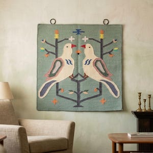 May include: A square wall hanging featuring a sage green background with two symmetrical cream-colored birds perched on stylized dark gray branches. The design includes small orange, yellow, and blue floral accents and two black metal rings at the top.