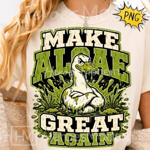 Könnte beinhalten: Ein cremefarbenes T-Shirt mit einer Grafik einer weißen Ente, die mit grünem Schleim bedeckt ist und die Arme verschränkt hat. Der Text "MAKE ALGAE GREAT AGAIN" steht in fetten, abgenutzten grünen Buchstaben, umgeben von Sumpfpflanzen und Schleimtropfen.