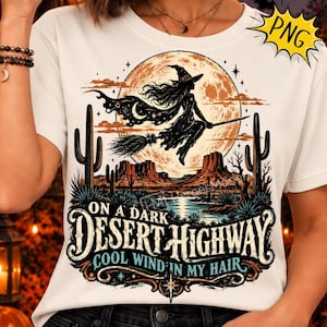 Puede incluir: Una camiseta blanca con un gráfico de una bruja volando en una escoba frente a una luna grande sobre un paisaje desértico con cactus y mesetas. El texto dice ON A DARK DESERT HIGHWAY COOL WIND IN MY HAIR en letras negras y azules.