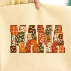 May include: A cream-colored sweatshirt featuring the word MAMA in large block letters. The letters are filled with a patchwork design of various autumn-themed fabrics in shades of orange, brown, green, and white, featuring floral and plaid patterns.