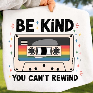 May include: White square fabric with the text "BE KIND YOU CAN'T REWIND" in black. A retro cassette tape design with a rainbow stripe is in the center. The design is surrounded by colorful star and sparkle accents.