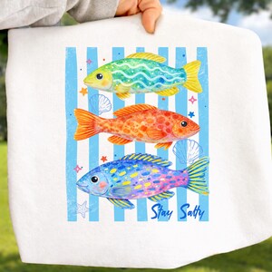 May include: White towel featuring three colorful fish illustrations against a blue and white striped background. The fish are green, orange, and blue, with yellow fins. The text "Stay Salty" is printed on the towel.