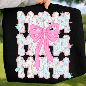 May include: Black tote bag featuring the word "MAMA" in a floral design, with a large pink bow. The letters are light blue with a white and floral design. Small flowers are scattered around the letters.