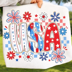 May include: A white t-shirt featuring a large graphic with the letters USA in a bold, stylized font. The letters are decorated with red, white, and blue patterns, including stars, stripes, and bows. The design is surrounded by red, white, and blue flowers.