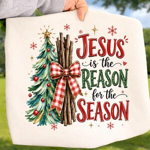May include: A white fabric item featuring a graphic of a green Christmas tree with red ornaments and a gold star, a bundle of brown sticks tied with a red and white checkered bow, and the text Jesus is the Reason for the Season.