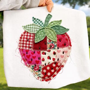 May include: A white square fabric with a large, patchwork strawberry design. The strawberry is composed of various fabric patterns, including red gingham, floral prints, and heart motifs. Green leaves and a checkered stem complete the design.